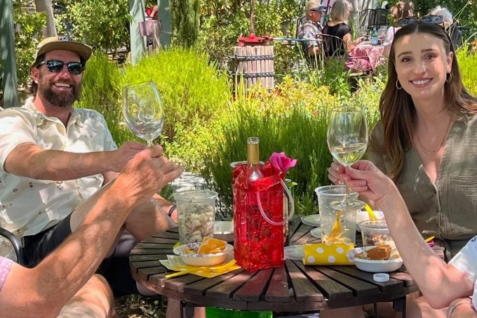 Two club members smiling holding glasses of our 2021 Pinot Grigio sitting at a table with a cooler bag holding the bottle of wine and plates of picnic food