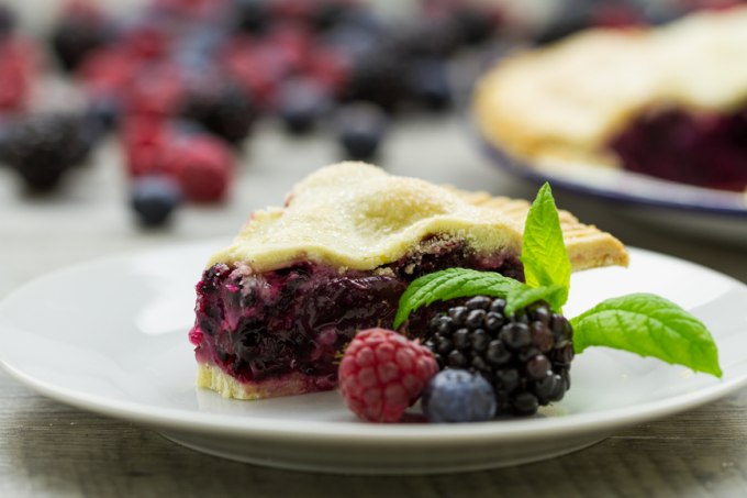 A close up of a slice of pie with top crust and filled with gooey blueberries and blackberries