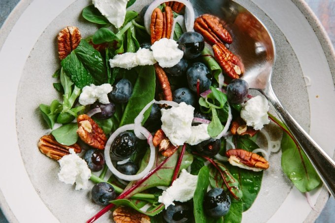 A white salad bowl filled with dark leafy greens, tossed with goat cheese chunks, nuts and vinaigrette dressing