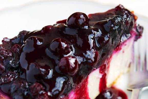 A slice of creamy cheesecake with a graham cracker crust topped with whole blueberry compote sauc