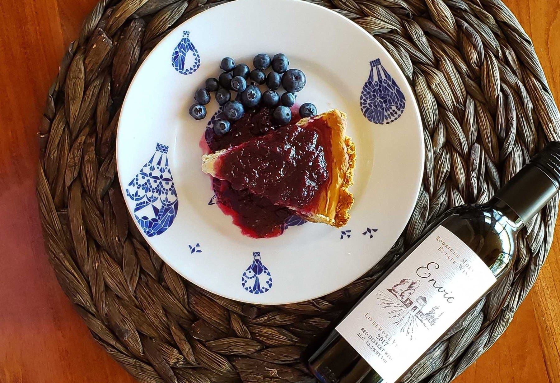 A slice of blueberry and blackberry pie sitting on a wooden table on a blue and white china plate with a bottle of RM 2017 Envie dessert wine next to it