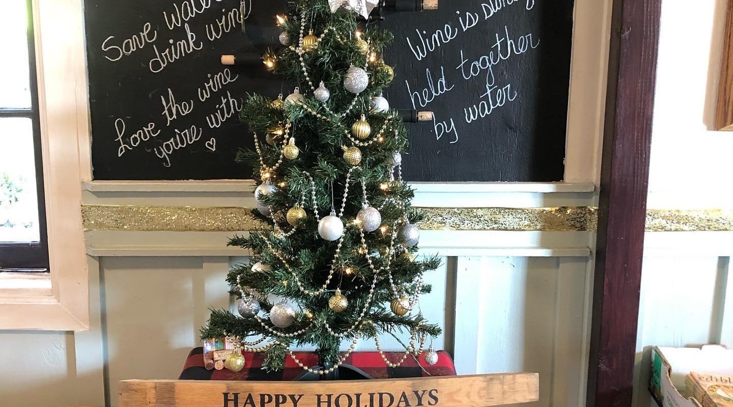 A small christmas tree decorated with white poinsettias sitting on top of a small electric fireplace in the tasting room with RM wine bottles hanging on the wall