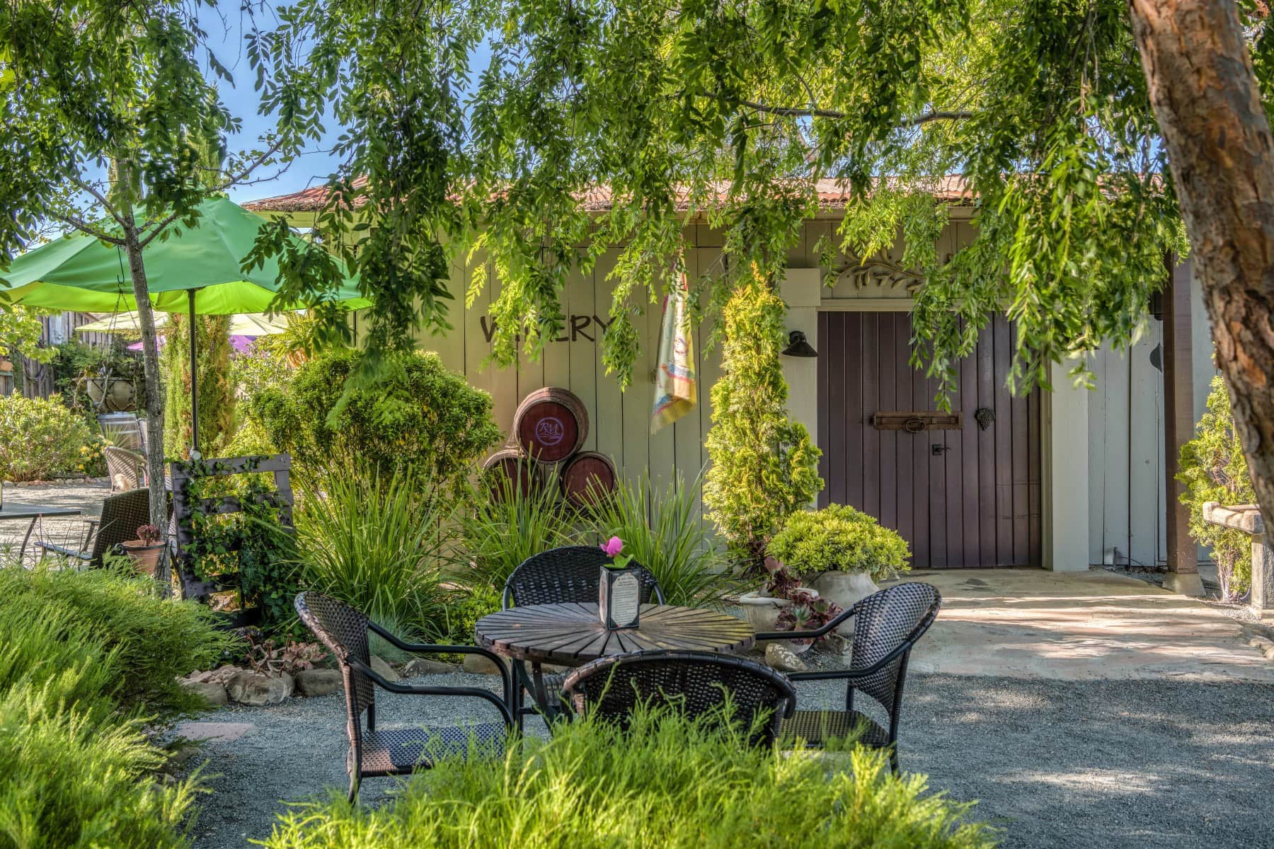 The tasting room in the background with an empty table in the foreground and a stack of 3 used oak barrels against the tasting room next to the front door