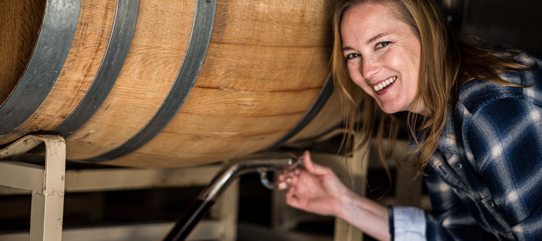 Winemaker Meredith Sarboraria is wearing a blue plaid shirt and smilingwhile using a wine thief to take a sample of wine straight from a barrel in the RM cellar
