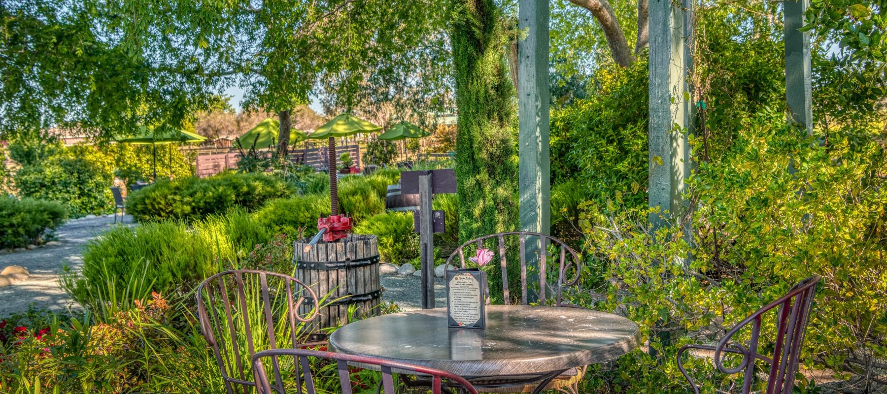 A table with four chairs in the RM wine tasting garden surrounded by green shrubs, trees, a trellis in the background and an old wooden wine press with a red handle