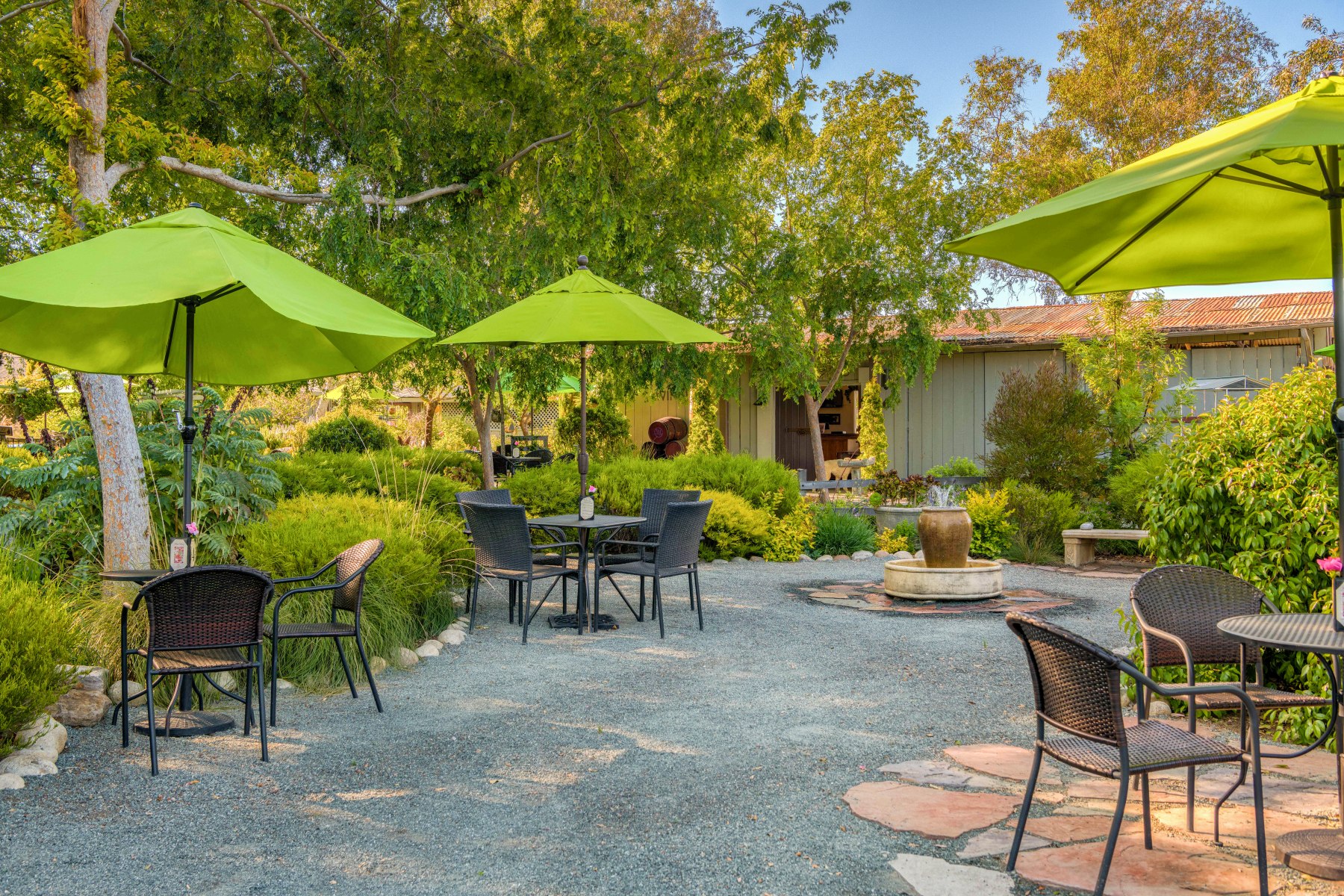 A tree lined garden with tables and umbrellas spaced around a centeral water foundtain